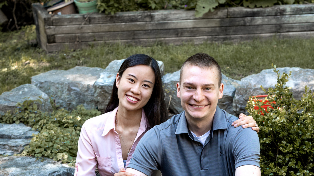 Wife and husband sitting on some rocks in the shape of heart with greenery in the background.