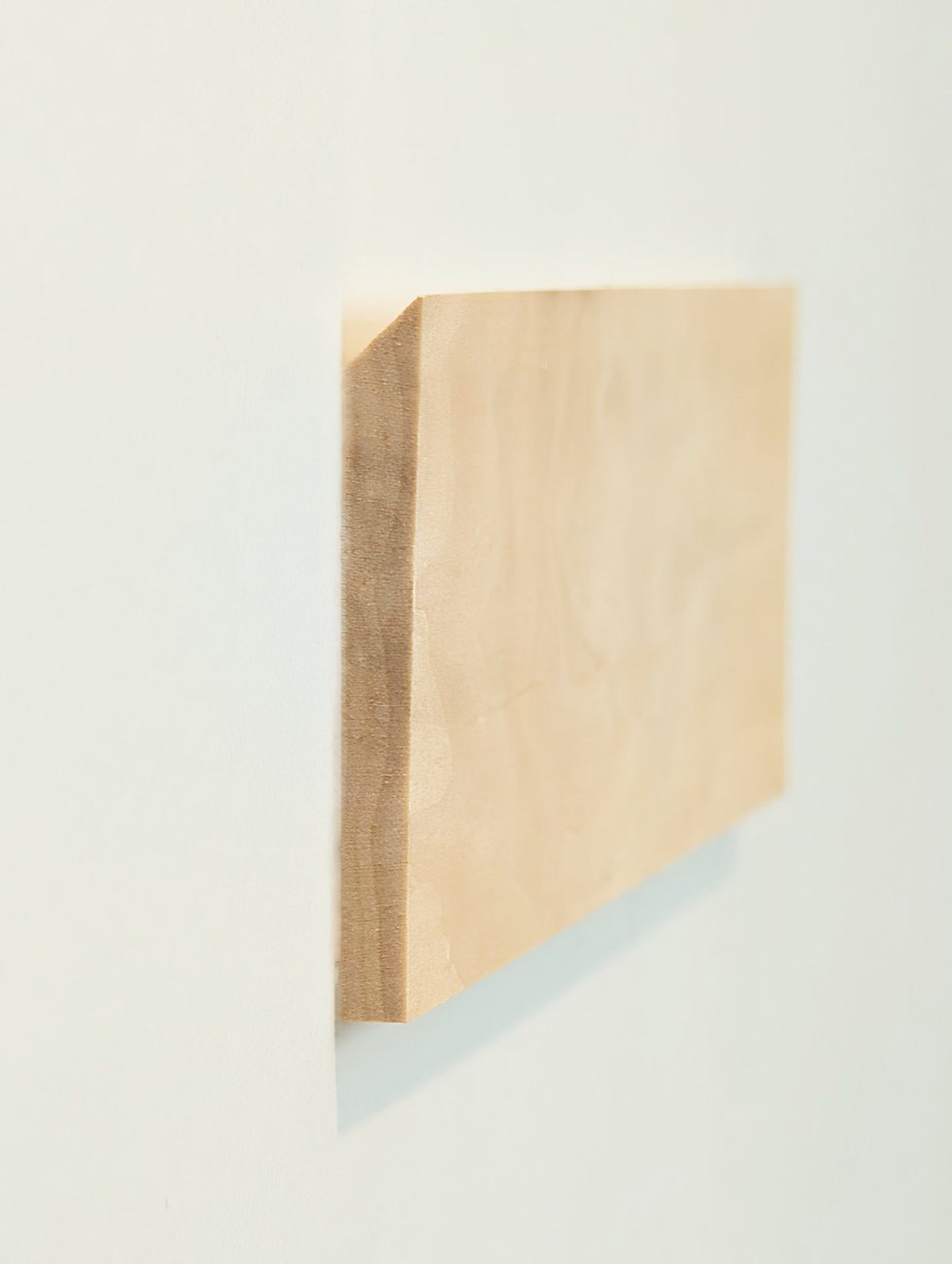 A rectangular light wood panel or board shown at an angle against a plain off-white wall, with visible grain and a slight shadow cast to the right.