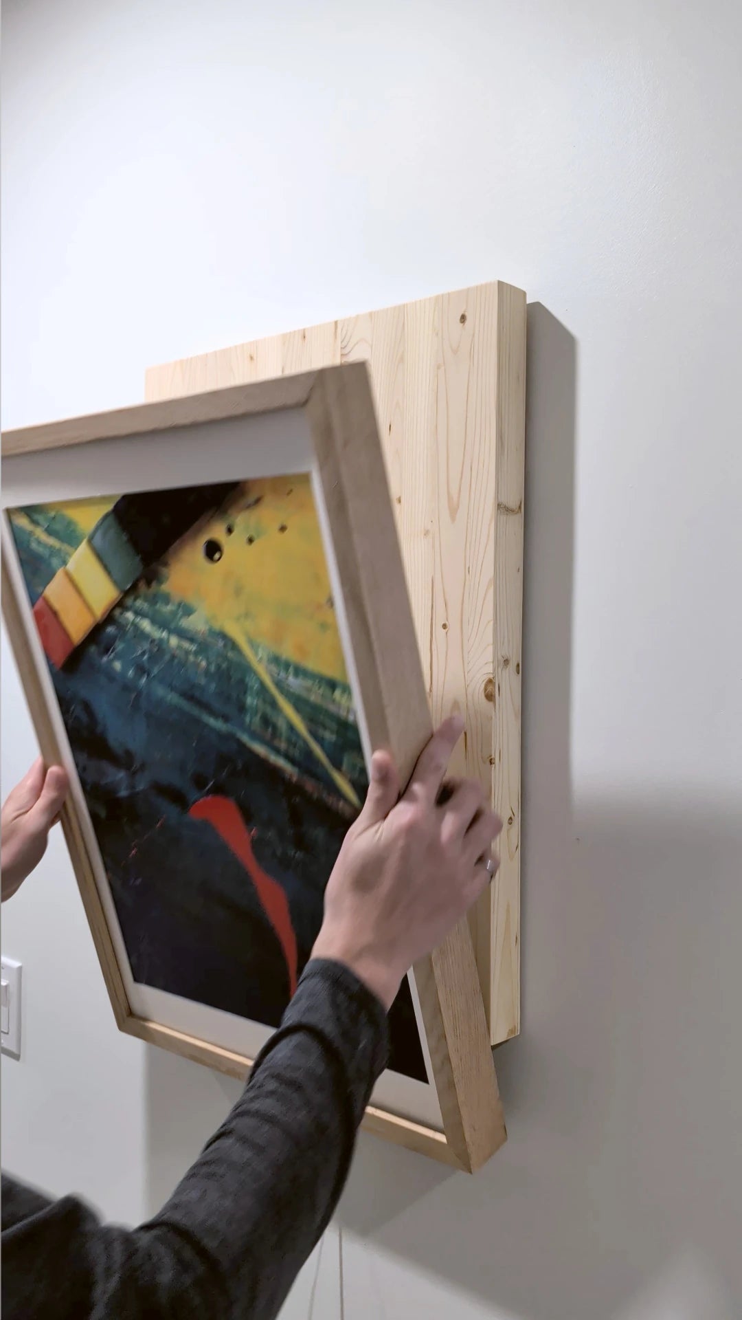 A person's hands are shown mounting a framed abstract art print onto a light-colored wooden wall-mounted frame against a plain white wall.